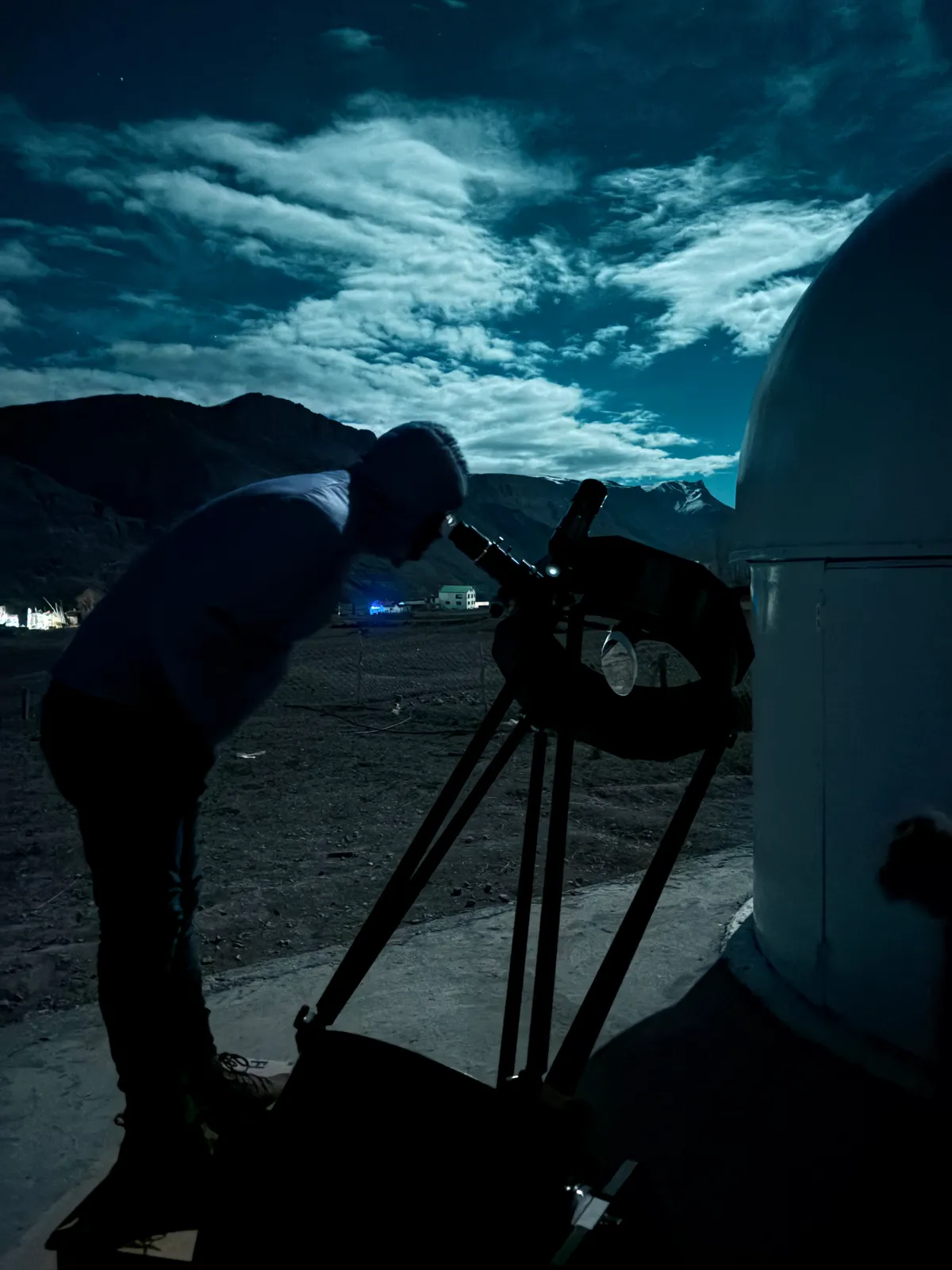Someone bent over a large telescope beside the AstroMonks dome under moonlit clouds