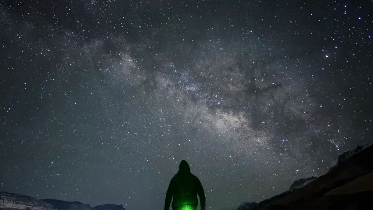 A person seen from behind, holding a green light under the Milky Way
